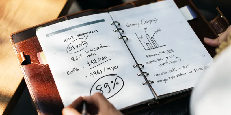 Une main note des calculs de coûts et de pourcentages dans un agenda.