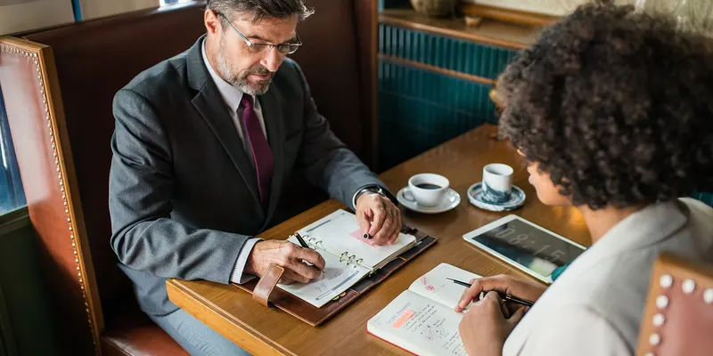Deux personnes échangent autour de documents, à une table de café.
